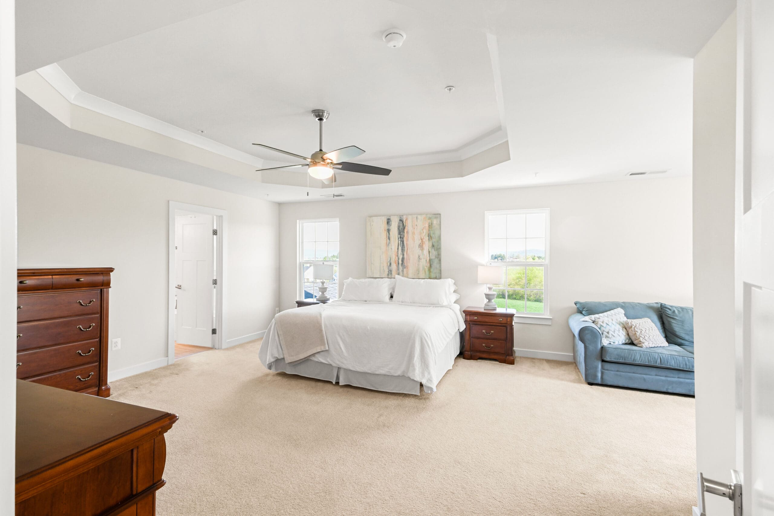 Large primary bedroom with tray ceiling, ceiling fan, neutral carpet, white bedding, and sitting area with blue loveseat and wood furniture